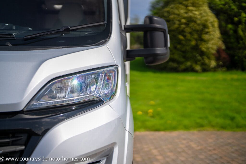 Close-up of the front left headlight and driver-side mirror of a white 2019 Swift Escape 694 Freestyle motorhome. A brick driveway and green lawn with bushes are visible in the background. The words "www.countrysidemotorhomes.com" are displayed in the bottom left corner of the image.