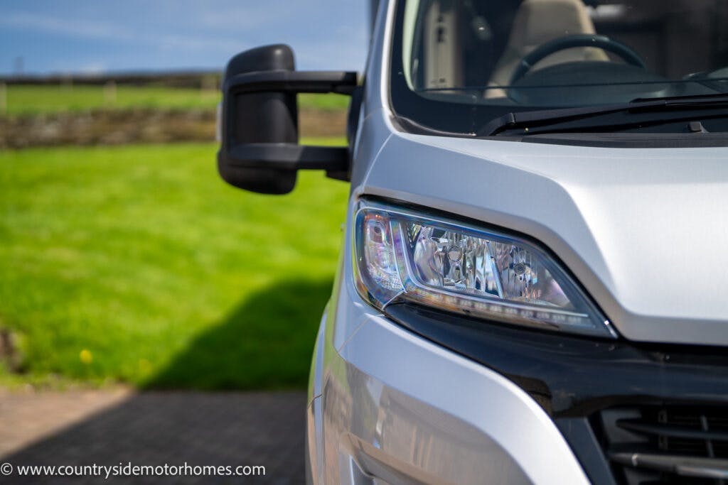 Close-up image of the front headlight and side mirror of a stationary white 2019 Swift Escape 694 Freestyle motorhome. The motorhome is parked on a paved area with a grassy field and a stone wall visible in the background. The URL www.countrysidemotorhomes.com is on the bottom left.