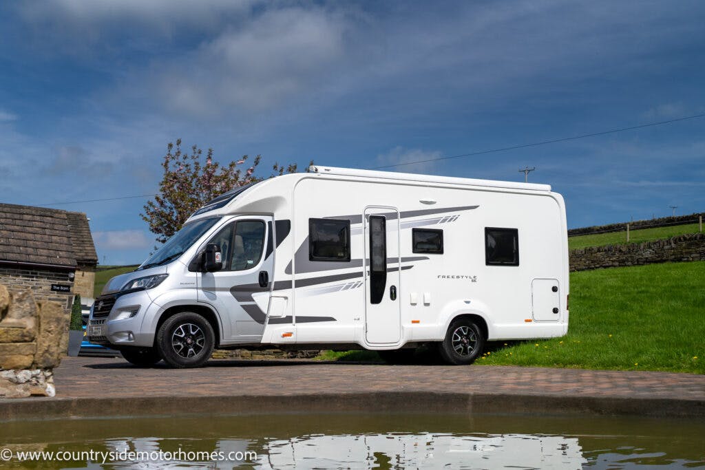 A 2019 Swift Escape 694 Freestyle motorhome is parked on a paved area near a grassy field and a stone building. The vehicle has dark-tinted windows and a sleek design with gray graphics on the side. The sky is partially cloudy with some blue patches visible.