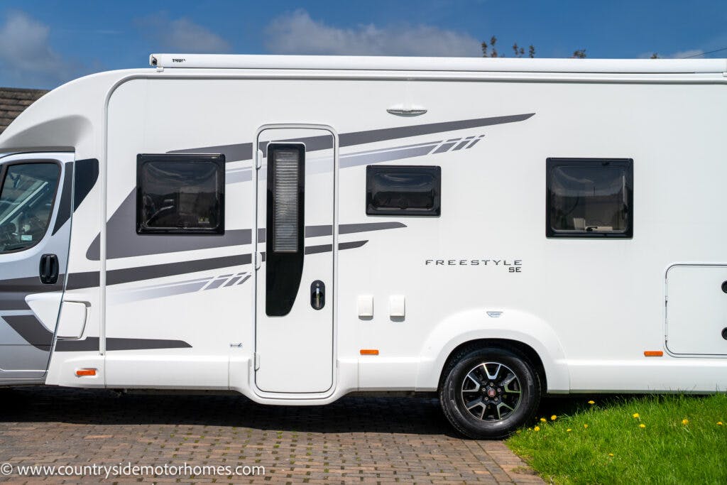 A side view of a modern white motorhome, the 2019 Swift Escape 694 Freestyle, parked on a paved surface. It features three windows and a central door with a black handle and frame. Abstract grey graphics adorn the side, with the motorhome brand "Freestyle SE" visible on the exterior.