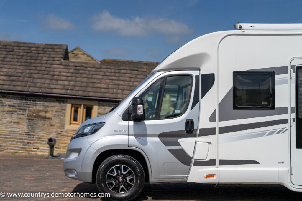 A white 2019 Swift Escape 694 Freestyle motorhome is parked on a paved surface in front of a rustic building with a tiled roof. The side of the motorhome features subtle grey graphics, perfectly complementing the serene countryside environment.