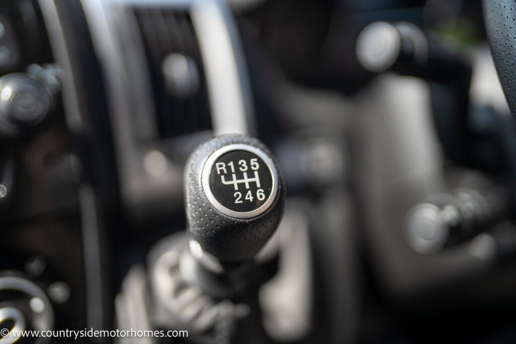 Close-up of the manual gear shift lever in a 2019 Swift Escape 694 Freestyle, labeled R 1 3 5 2 4 6. The background reveals various parts of the car's dashboard and controls. The image URL, www.countrysidemotorhomes.com, is visible in the bottom left corner.