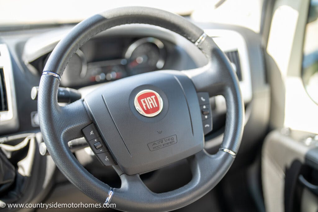 Close-up image of a Fiat car steering wheel with control buttons and the company logo in the center. The dashboard and part of the car interior are visible in the background, showcasing the elegance of the 2019 Swift Escape 694 Freestyle. The image also includes "www.countrysidemotorhomes.com" in the bottom left corner.