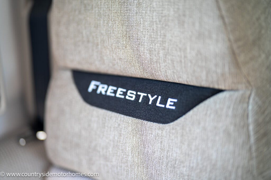 Close-up of a car seat labeled "FREESTYLE" on the backrest, with beige upholstery and black fabric in the middle displaying the label. The website "www.countrysidemotorhomes.com" is visible in the bottom left corner, showcasing details of the 2019 Swift Escape 694 Freestyle.