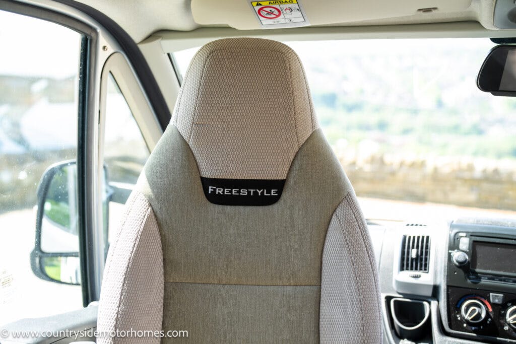 A close-up view of the driver's seat inside a 2019 Swift Escape 694 Freestyle motorhome, featuring light beige and gray upholstery with the word "FREESTYLE" embroidered on the headrest. The interior of the motorhome, including part of the dashboard and steering wheel, is visible.