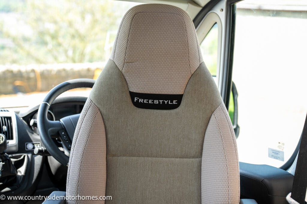 A driver's seat inside a 2019 Swift Escape 694 Freestyle vehicle, labeled "Freestyle" on the headrest. The seat is upholstered in a combination of light tan and gray fabric. The steering wheel, dashboard, and a window are partially visible in the background.