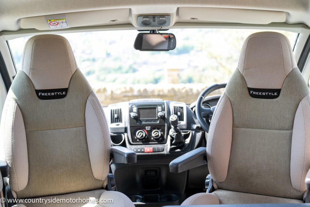 The interior of the 2019 Swift Escape 694 Freestyle motorhome features two front seats labeled "Freestyle" and a central dashboard with controls. Through the windshield, a blurry outdoor scene is visible. The image has a watermark at the bottom left corner and a label on the ceiling.