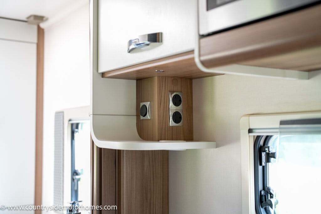 Close-up of the interior of a 2019 Swift Escape 694 Freestyle camper van kitchen area, showing a wooden shelf with electrical outlets, cabinetry above, and part of a window to the right. The image highlights the functionality and design of the space. The website address is visible in the bottom-left corner.