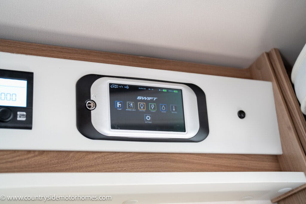 A touchscreen control panel for the 2019 Swift Escape 694 Freestyle motorhome is mounted on a wooden surface. The display shows various icons and options for controlling the motorhome's systems. A small screen to the left shows a numerical display.