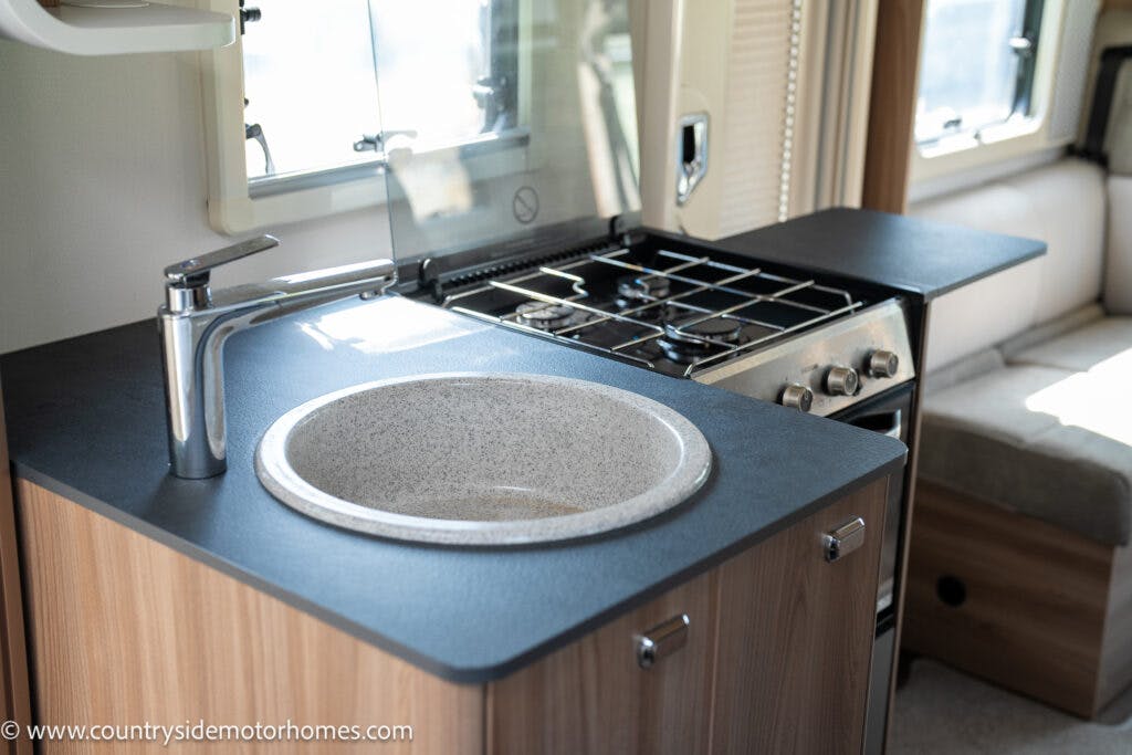 A modern RV kitchen in the 2019 Swift Escape 694 Freestyle features a round sink with a sleek faucet, situated on a black countertop. To the right, there is a stainless steel gas stove and oven. A window with blinds is located above the counter, and part of a cushioned seating area is visible.