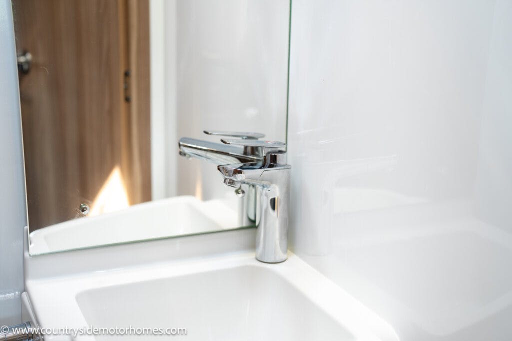 A shiny chrome faucet is mounted on a white sink, with a mirrored backsplash directly behind it. The faucet handle is positioned to the right. The surrounding area is clean, reminiscent of the pristine interiors found in the 2019 Swift Escape 694 Freestyle, with a wooden door partially visible in the background.