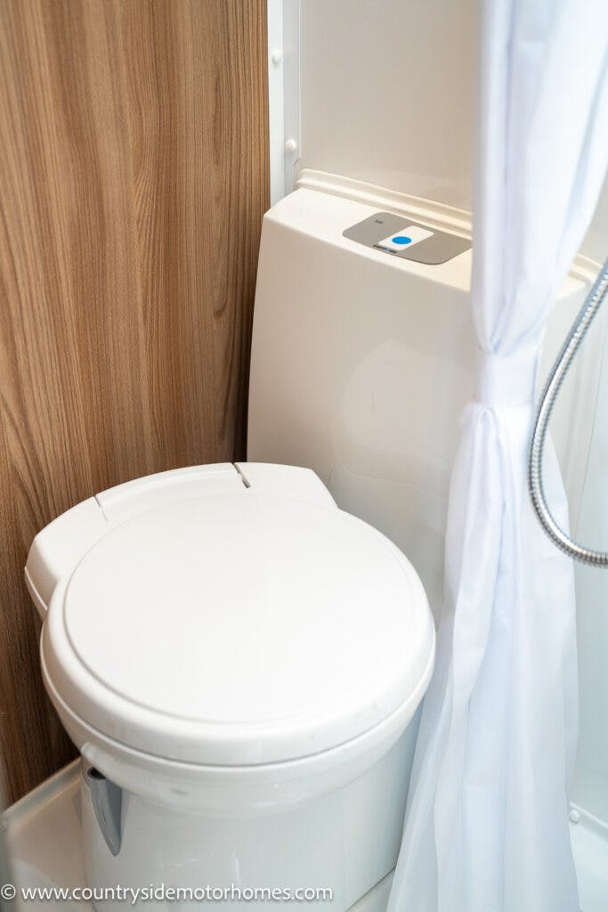 The image shows a white toilet with a closed lid positioned near a wooden panel. A white shower curtain hangs to the right of the toilet. The compact, clean bathroom suggests this could be part of the 2019 Swift Escape 694 Freestyle RV or a small living space.