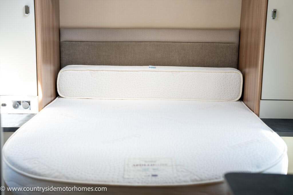The image shows a neatly made bed with a white mattress inside what appears to be a 2019 Swift Escape 694 Freestyle motorhome. The headboard is cushioned, and there are wooden cabinets on either side. Natural light streams in from the left side of the image.