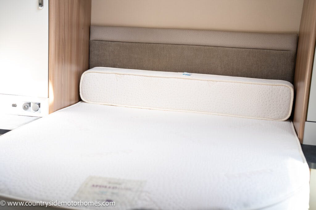A neatly made bed in the 2019 Swift Escape 694 Freestyle motorhome, featuring a beige headboard and a mattress with a white, textured cover. The setting includes light wood paneling and built-in storage, providing a compact and cozy sleeping area.