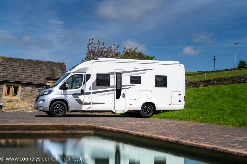 The 2019 Swift Escape 694 Freestyle motorhome is parked on a paved area near a house with a stone exterior. It sits adjacent to a pond, in front of a green field under a partly cloudy sky. Visit www.countrysidemotorhomes.com for more details.