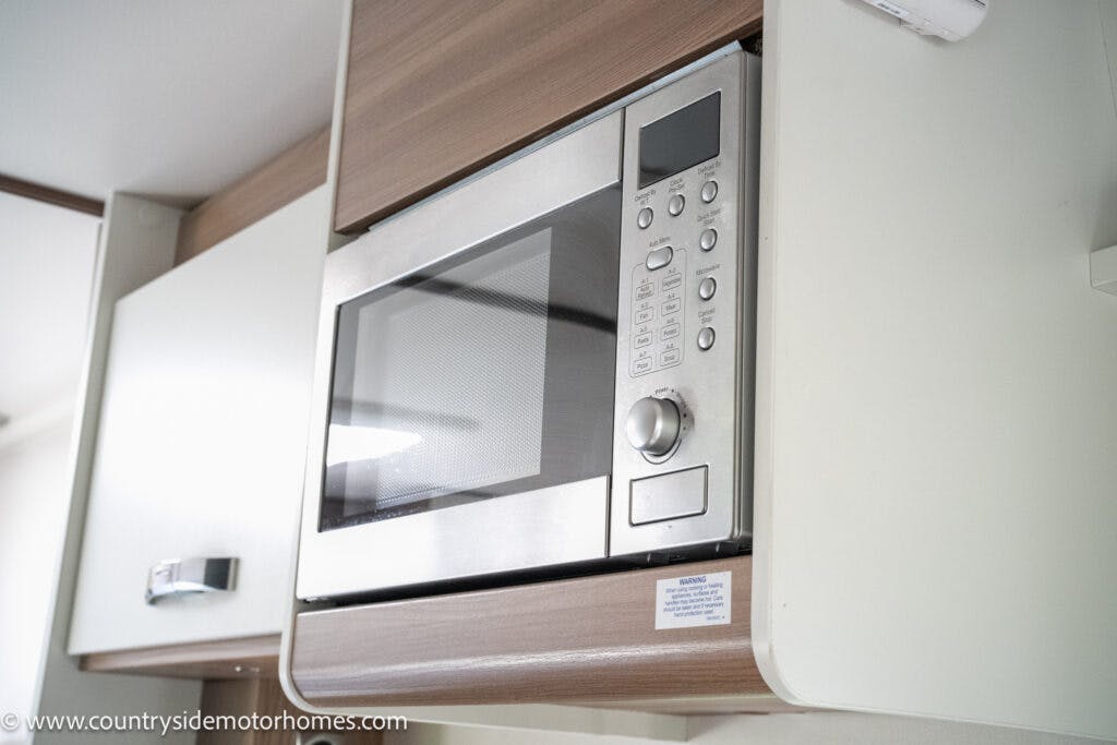 A stainless steel microwave is installed in a wooden cabinet, mounted above a white cabinet in the kitchen area of a 2019 Swift Escape 694 Freestyle. Various buttons and a digital display are visible on the microwave. The website "www.countrysidemotorhomes.com" is visible at the lower-left corner of the image.