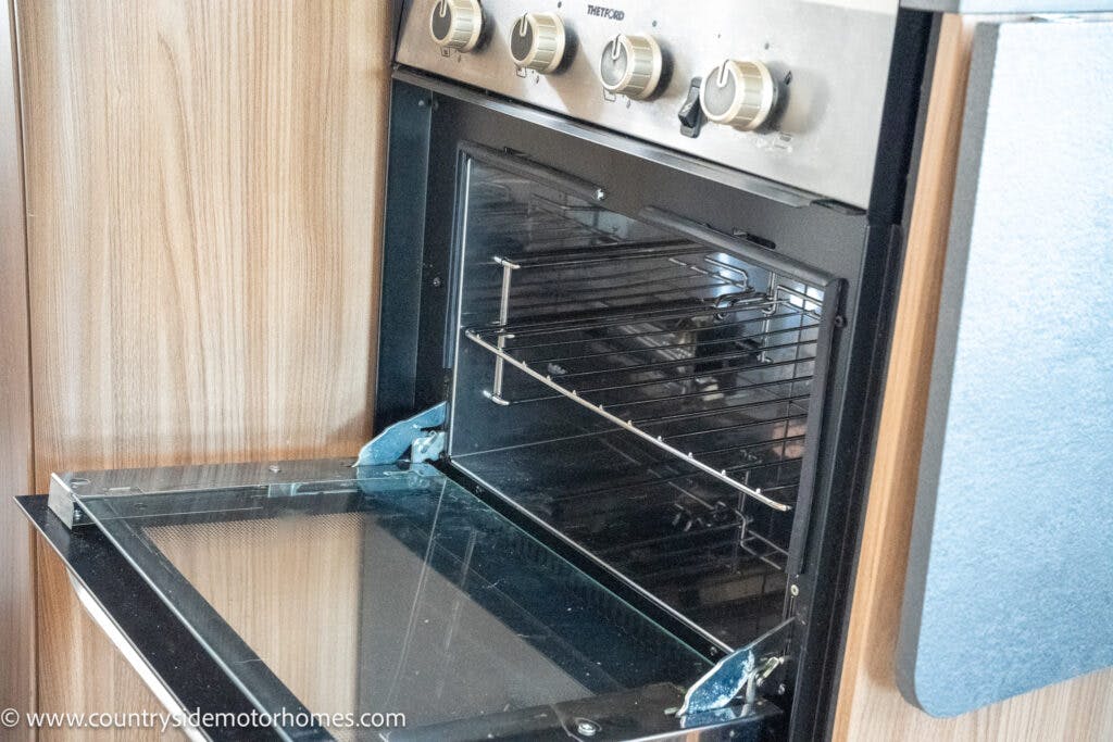 A close-up image of an open stainless steel oven with a glass door in the 2019 Swift Escape 694 Freestyle. The oven boasts multiple metallic dials on the control panel and is set into a wooden cabinet. Inside, wire racks and a rotisserie attachment are visible, showcasing its versatile cooking capabilities.