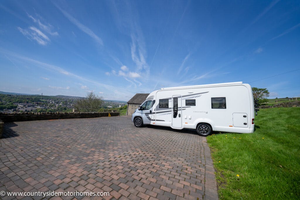 A 2019 Swift Escape 694 Freestyle motorhome is parked on a large, paved driveway with a scenic hilly landscape in the background. The sky is clear with a few wispy clouds. The driveway is bordered by a grassy area on one side. Visit www.countrysidemotorhomes.com for more details.