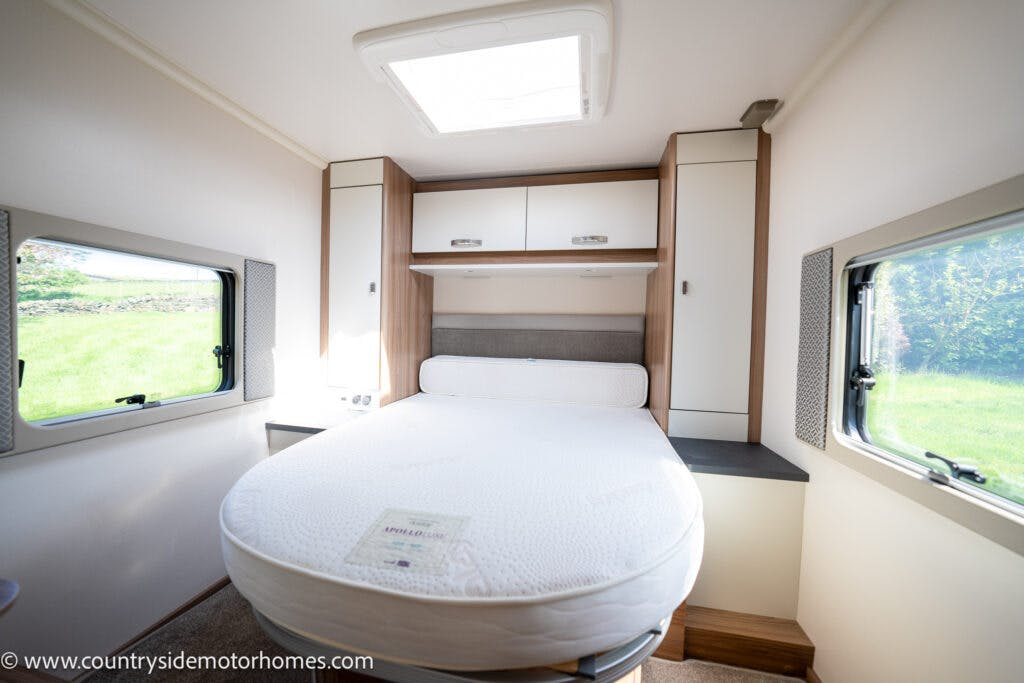 The image shows the interior of a 2019 Swift Escape 694 Freestyle motorhome bedroom with a round mattress on a bed frame against the back wall. There are cabinets above and beside the bed, two windows on either side, and a skylight in the ceiling. The walls and furniture are in light colors.