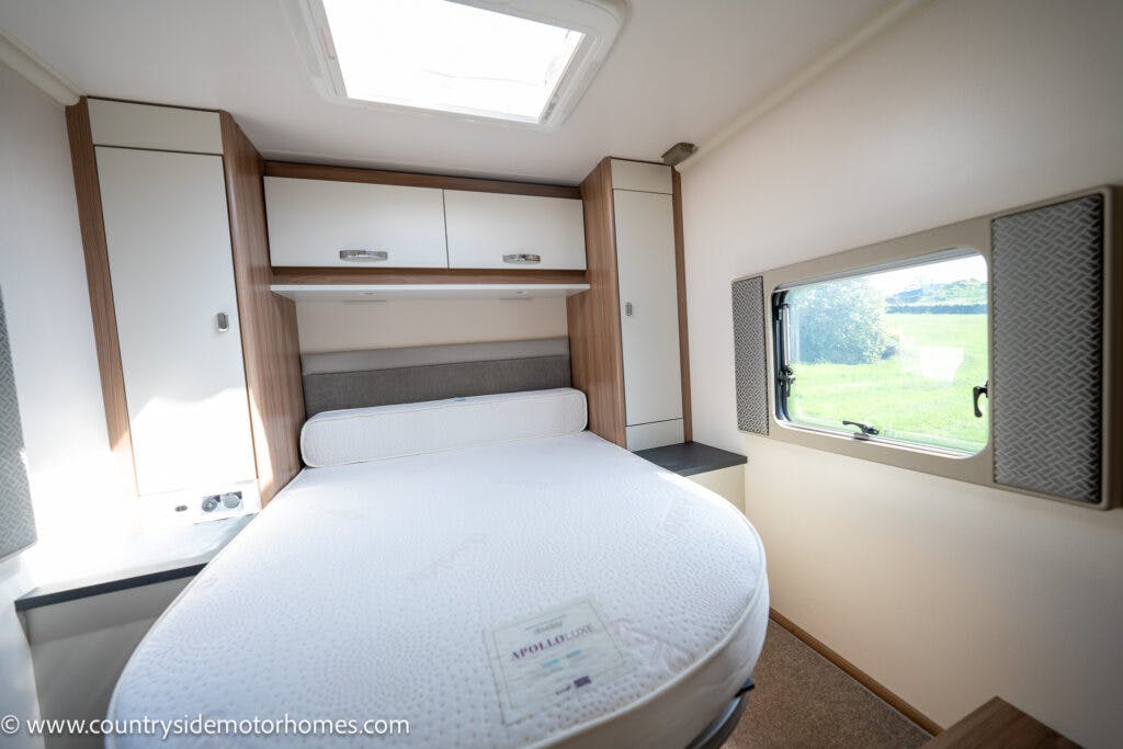 A compact, well-lit bedroom inside the 2019 Swift Escape 694 Freestyle motorhome features a round mattress with a headboard underneath storage cabinets. A small window is present on the right wall, and a skylight above provides additional natural light.