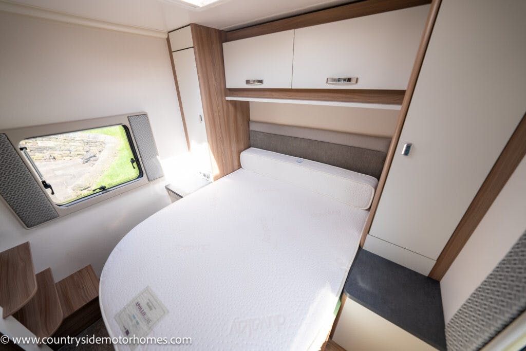The interior of the 2019 Swift Escape 694 Freestyle motorhome bedroom features a corner-mounted bed with a white mattress, light wooden and white cabinetry above, and a small window to the left. The floor has a step-up design, and there is a gray and black bench beside the bed.