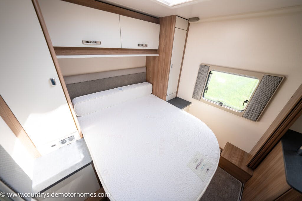 Interior of a 2019 Swift Escape 694 Freestyle motorhome bedroom featuring a fitted double bed against a wall, with under mounted storage cabinets, a small window with curtains, and a wardrobe cabinet on the side. The mattress is uncovered, and natural light streams through the window.