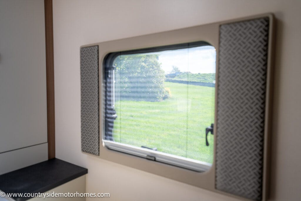 The image shows the interior of a 2019 Swift Escape 694 Freestyle motorhome from Countryside Motorhomes, focusing on a window with a view of a green field and trees outside. The window is framed with patterned panels on either side. The web address is visible in the bottom-left corner.