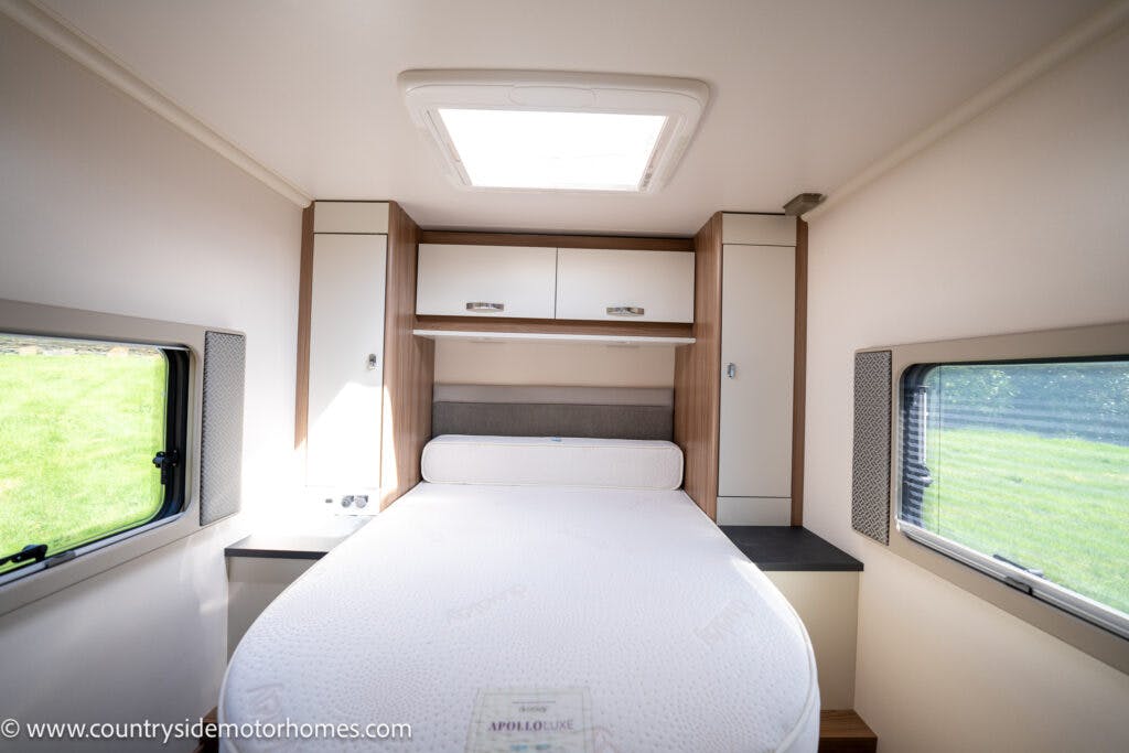 The image showcases the interior of a 2019 Swift Escape 694 Freestyle motorhome bedroom, featuring a double bed with a white mattress. Wooden cabinets are mounted above for storage. Natural light streams through windows on both sides and a skylight above. The bed includes a roll cushion at the head.