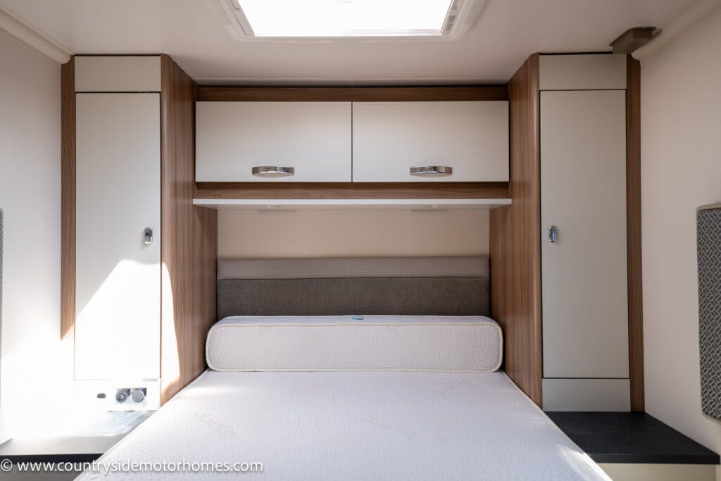 The image shows the interior of a 2019 Swift Escape 694 Freestyle motorhome bedroom with a compact, neatly made bed. Above the bed are overhead storage cabinets with light wood and white finishes. The room is well-lit, and there are additional vertical storage units on either side of the bed.