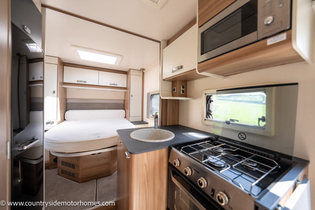 Interior of a 2019 Swift Escape 694 Freestyle motorhome with a kitchenette and sleeping area. The kitchenette includes a stove, oven, microwave, sink, and cabinets. The sleeping area features a bed with storage compartments. Bright natural light shines through the windows and skylight.