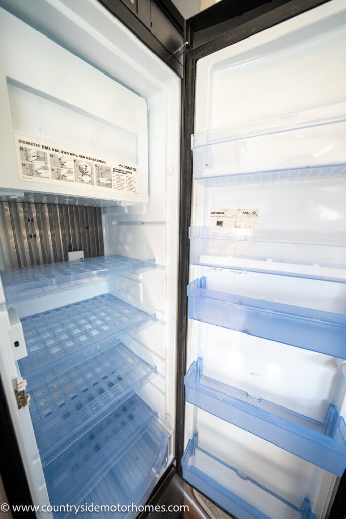An open refrigerator with a top freezer compartment and an empty main section inside a 2019 Swift Escape 694 Freestyle. The inside features multiple clear shelves and door racks. The refrigerator is clean and well-lit, displaying a Dometic brand label. A website URL is visible at the bottom of the image.