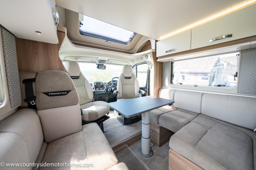 Interior of the 2019 Swift Escape 694 Freestyle motorhome featuring a seating area with light brown upholstery, a square table, and a driver's cab visible in the background. Large windows provide natural light. The overall design is modern and cozy.