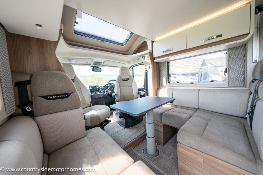 The interior of a 2019 Swift Escape 694 Freestyle motorhome is shown. It features beige and brown seating, a gray rectangular table, and storage cabinets. A window at the front provides natural light. The website www.countrysidemotorhomes.com is visible in the lower left corner.