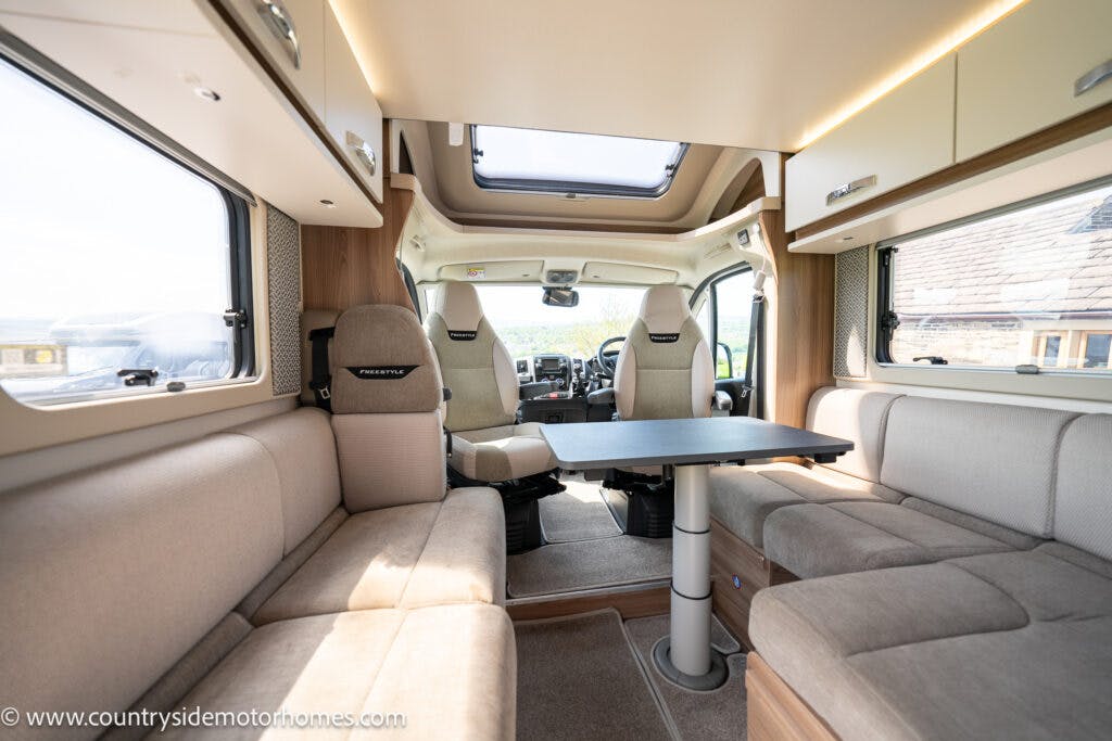 The interior cabin of the 2019 Swift Escape 694 Freestyle motorhome features a seating area with beige cushions, a central foldable table, and driver and passenger seats at the front. Large windows provide ample natural light. Logo "Prestige" visible on the seats. Website URL in corner.