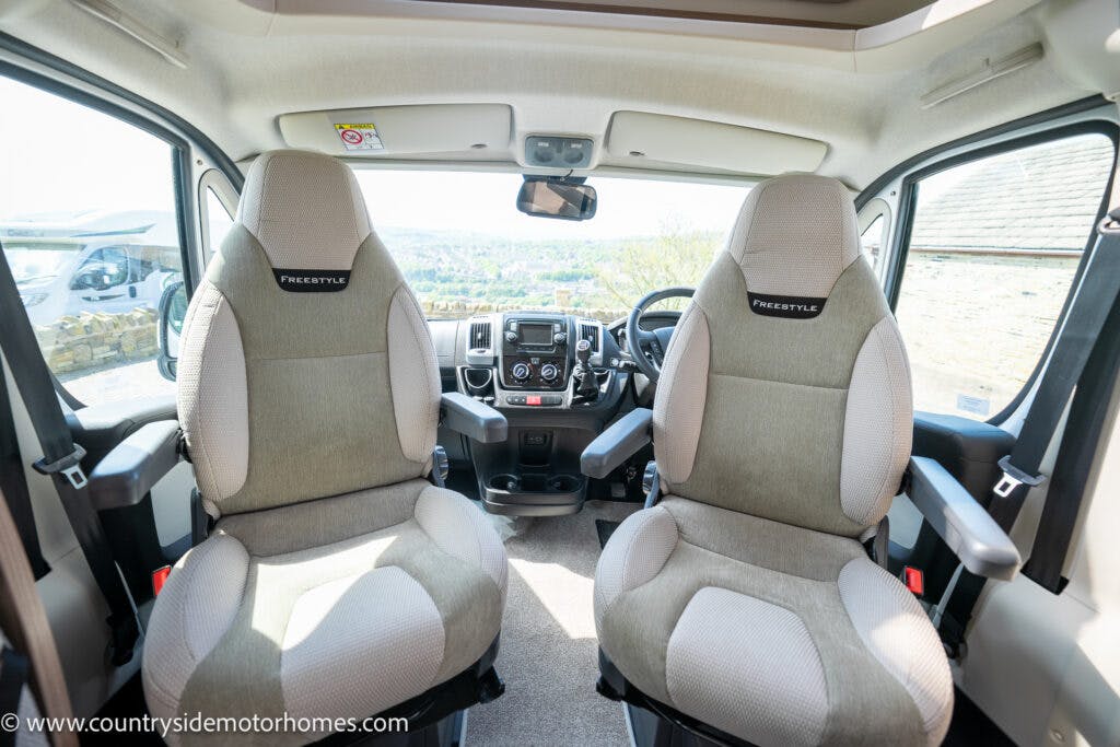 This image shows the interior of a 2019 Swift Escape 694 Freestyle motorhome's driver's cabin. It features two beige captain's chairs labeled "FreeStyle," a center console with a steering wheel and controls, large windows, and a panoramic view. The URL "www.countrysidemotorhomes.com" is visible.