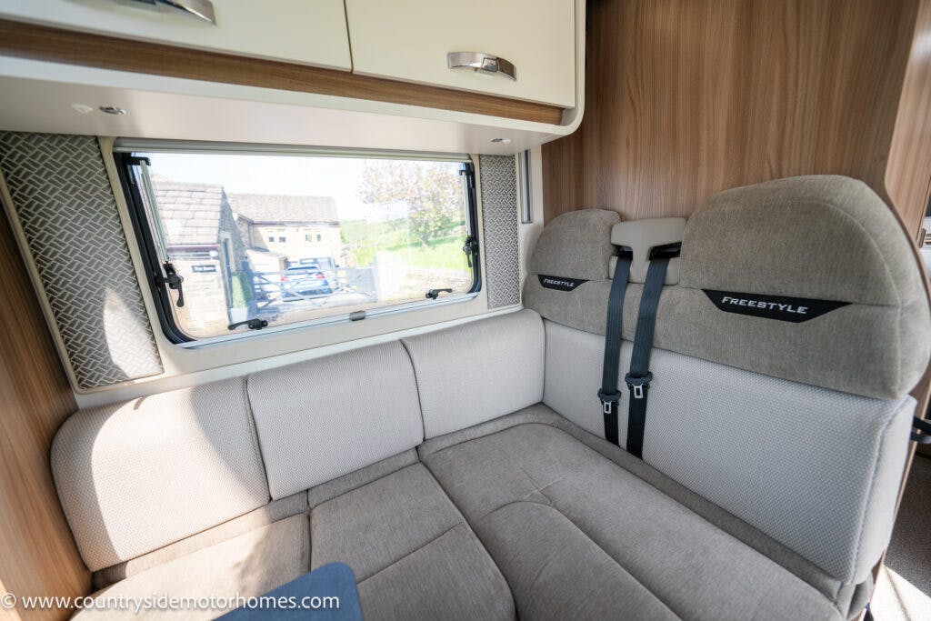 The image shows the interior of a 2019 Swift Escape 694 Freestyle motorhome with a seating area. The seats are upholstered in gray fabric with "Freestyle" embroidered on them. There is a window above the seating area and a wooden cabinet above the window. Website URL: www.countrysidemotorhomes.com.