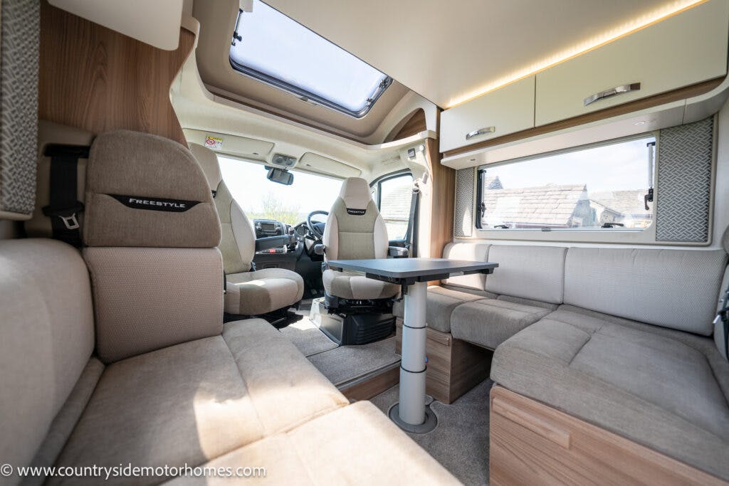 The image shows the interior of a 2019 Swift Escape 694 Freestyle motorhome with beige and gray upholstery. It features a seating area with a table in the center, large windows, and overhead storage cabinets. A skylight is visible above, and the website www.countrysidemotorhomes.com is in the bottom left corner.
