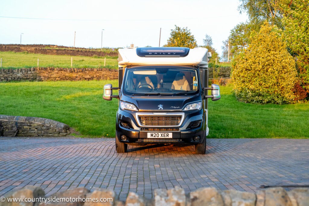A black Peugeot campervan with a "Bailey" logo and the model name 2021 Bailey Autograph 79-4I is parked on a paved driveway. In the background are a stone wall, lush green grass, and trees. The vehicle's license plate reads "M20 XER." The website "countrysidemotorhomes.com" is visible in the bottom-left corner