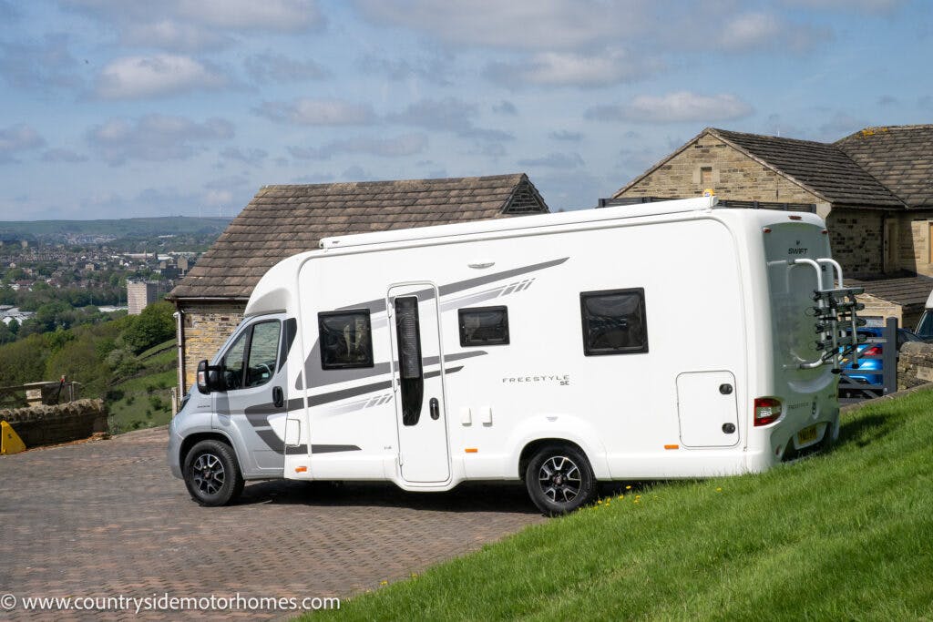 A 2019 Swift Escape 694 Freestyle motorhome with gray decals is parked on a brick driveway in front of a stone building. The surrounding landscape includes green grass, distant trees, and a view of a town. The motorhome has windows and a side door.