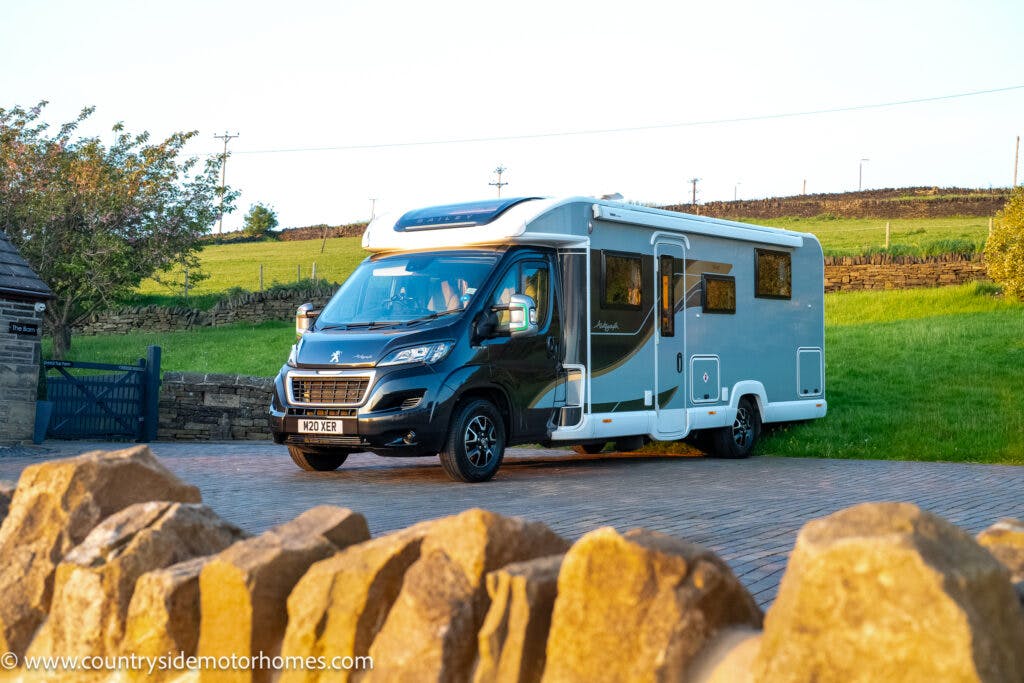 A 2021 Bailey Autograph 79-4I motorhome is parked on a paved driveway in a rural setting. The vehicle has a two-tone design with a black front and white rear. There is a stone wall in the foreground and a grassy field with a wire fence in the background.