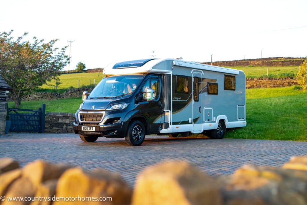 A 2021 Bailey Autograph 79-4I motorhome parked on a paved driveway. The vehicle is primarily black with white and grey accents. The background features a grassy area with a stone fence and hills, creating a serene countryside setting complete with trees and a clear sky.