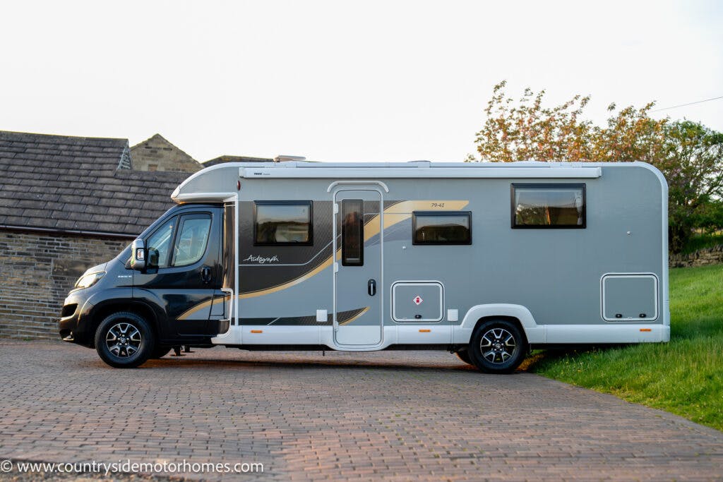 A grey and white 2021 Bailey Autograph 79-4I is parked on a brick driveway next to a house with a slate roof. The motorhome features a streamlined design and is equipped with multiple windows, a door, and external storage compartments.