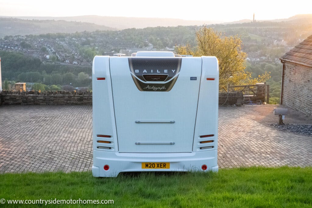The rear view of a white 2021 Bailey Autograph 79-4I motorhome parked on a paved surface with a scenic view of hills and a small village in the background. The license plate reads M20 XER. A URL for countrysidemotorhomes.com is visible in the bottom left corner.