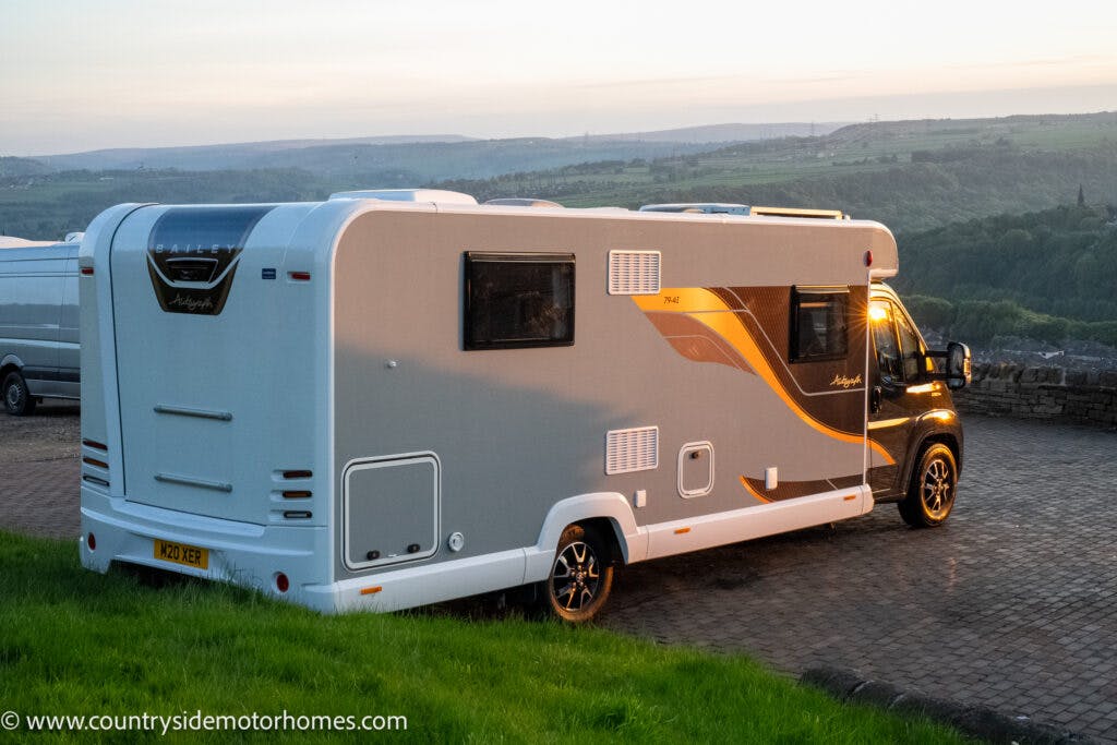 A 2021 Bailey Autograph 79-4I motorhome is parked on a paved area. It features a sleek design with grey and orange accents. The surrounding area is scenic with lush green fields and hills in the background. The evening light casts a warm glow on the vehicle.