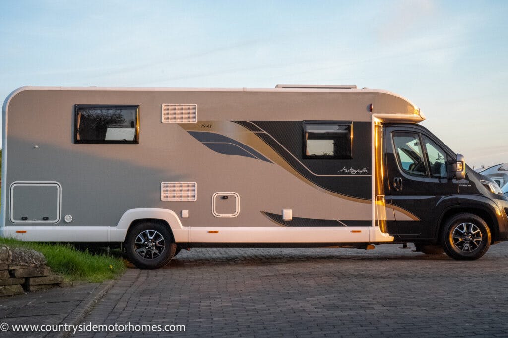 A grey and white 2021 Bailey Autograph 79-4I motorhome with black accents is parked on a paved surface. It has multiple side windows, one of which is open, and a lit entry door. The vehicle features a black cab and alloy wheels. The website "www.countrysidemotorhomes.com" is visible in the bottom left corner.