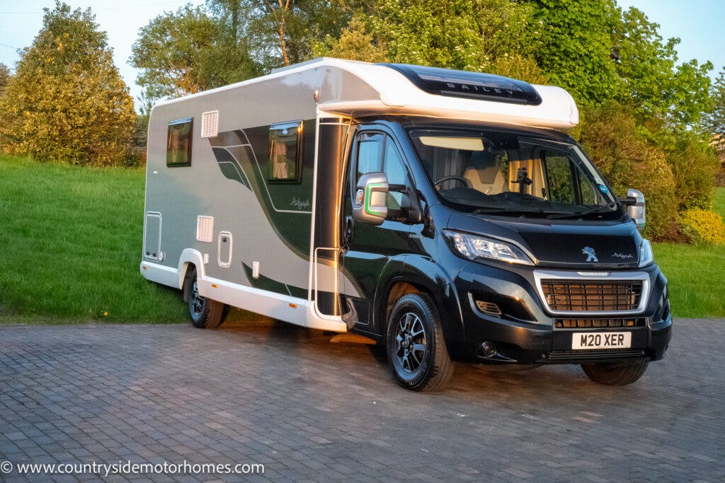 A 2021 Bailey Autograph 79-4i motorhome with a Peugeot chassis is parked on a paved area with a grassy background. The vehicle boasts a black front and greenish-gray sides with white accents. The website "www.countrysidemotorhomes.com" is displayed at the bottom left of the image.