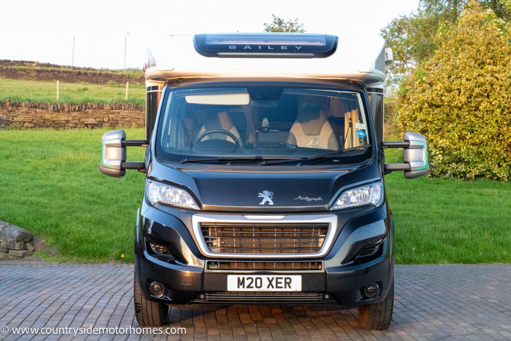 A black and white 2021 Bailey Autograph 79-4i motorhome with the word "Bailey" on the top front and a Peugeot logo below it is parked on a brick driveway. It has side mirrors extended and a license plate reading "M20 XER." The background includes a grassy area and a brick wall.