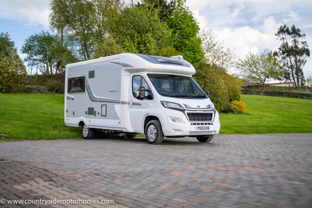 A white 2021 Auto-Sleepers Broadway EL motorhome is parked on a brick-paved driveway in front of a grassy area with trees. The motorhome has the license plate PX21 EXW and displays the logo "Autograph" on its side. A website URL is visible in the bottom left corner of the image.