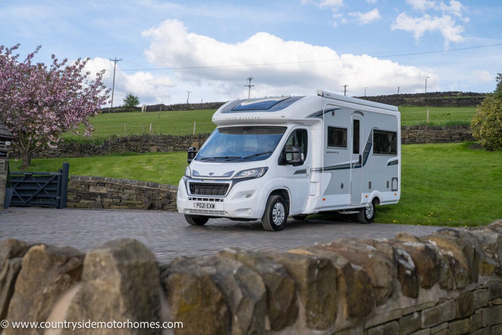A 2021 Auto-Sleepers Broadway EL motorhome is parked on a cobblestone driveway in a rural area. The sky is partly cloudy, and there is a green field and stone walls in the background. A flowering tree is visible on the left. The motorhome has the website www.countrysidemotorhomes.com on it.