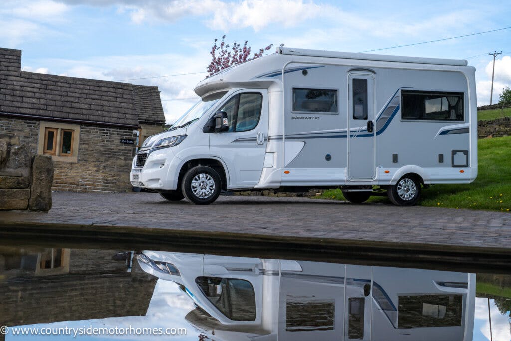 A white 2021 Auto-Sleepers Broadway EL motorhome is parked on a paved driveway next to a brick building. The reflection of the motorhome is visible in a body of water in the foreground. The sky is partly cloudy, and there is green grass in the background.